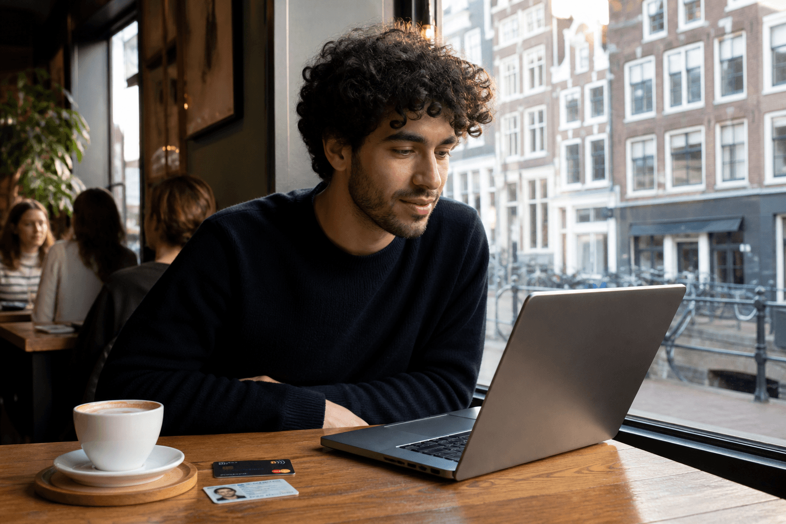 Photorealistic Dutch student café table: multicultural student reviewing a laptop enrolment portal beside a debit card, student ID card and coffee, soft canal-house daylight through the window.